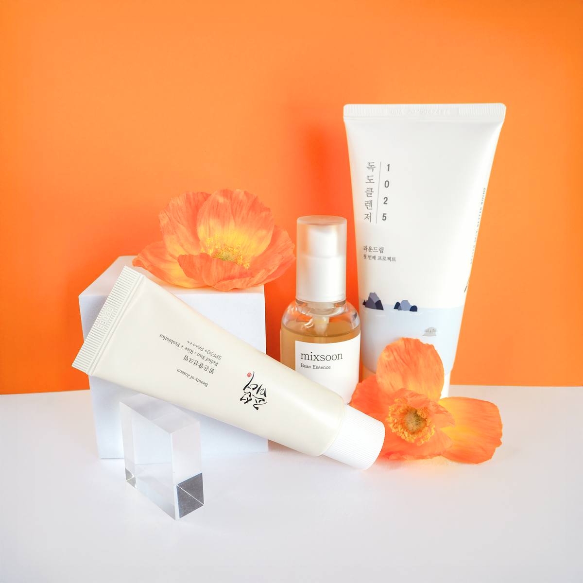 Auf einem Orangen Hintergrund drei Produkten - Cleanser, Bean Essence, Sonneschutzcreme. Die Sonnenschutzcreme ist auf einem Glas-Rechteck angelehnt und es gibt zwei große Orangene Blumen auch als Teil der Komposition.