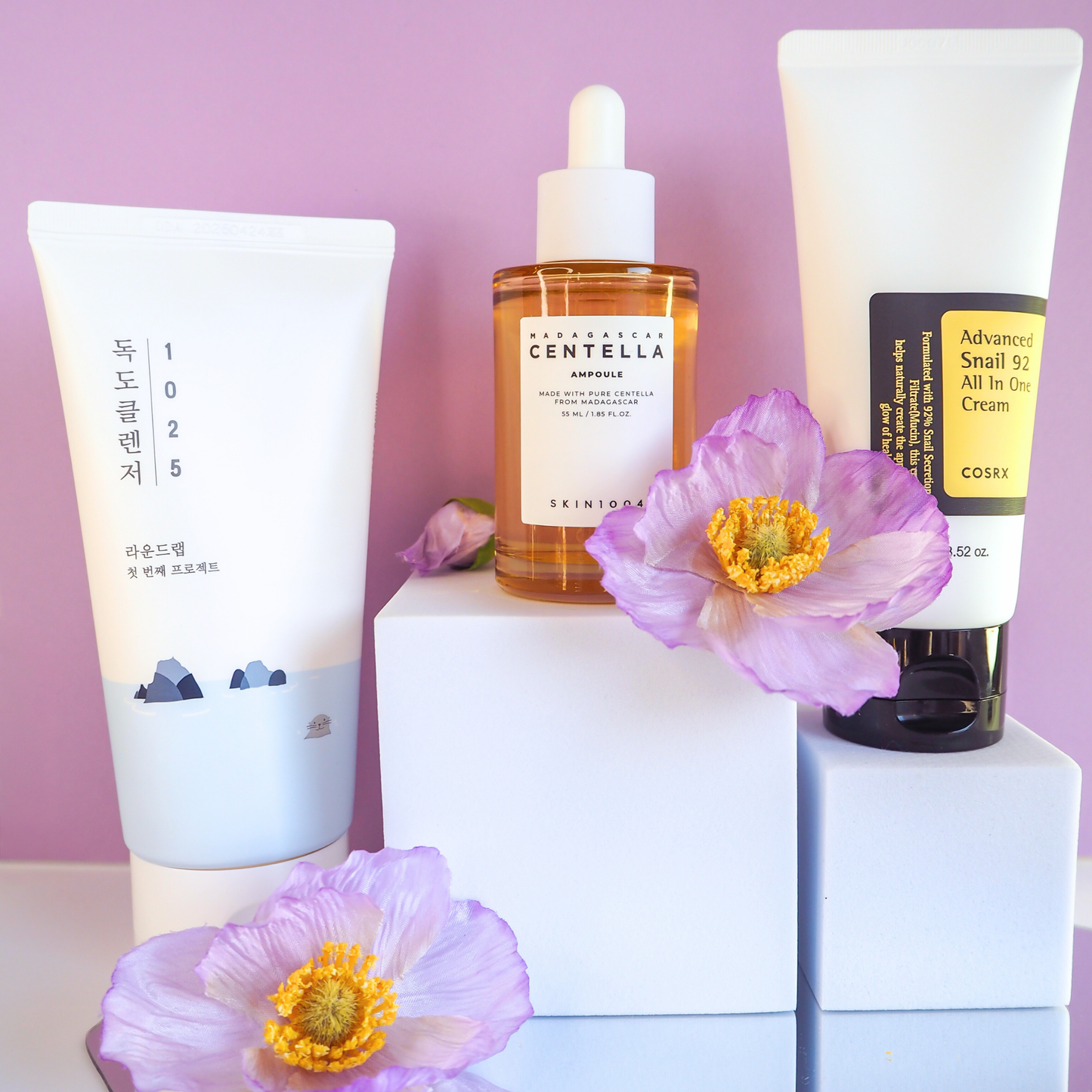 K-Beauty Produkte auf weißen Podesten vor lila Hintergrund mit dekorativen Blüten: Round Lab 1025 Cleanser, SKIN1004 Centella Ampoule und COSRX Snail 92 Creme