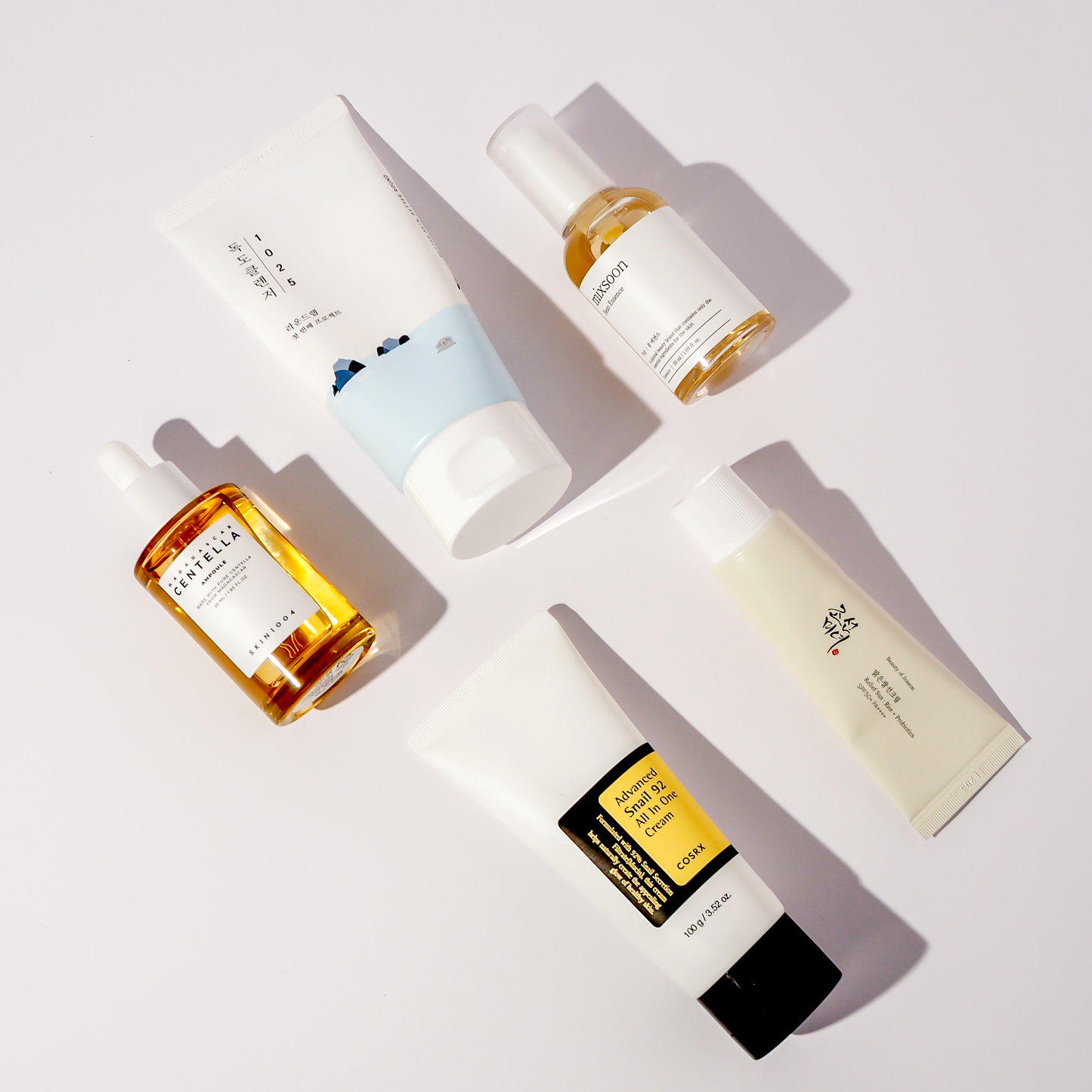Flatlay von fünf K-Beauty Produkten: Round Lab 1025 Dokdo Creme, mixsoon Bean Essence, SKIN1004 Madagascar Centella Ampoule, COSRX Advanced Snail 92 All In One Cream und Beauty of Joseon Relief Sun Cream SPF50+ PA++++