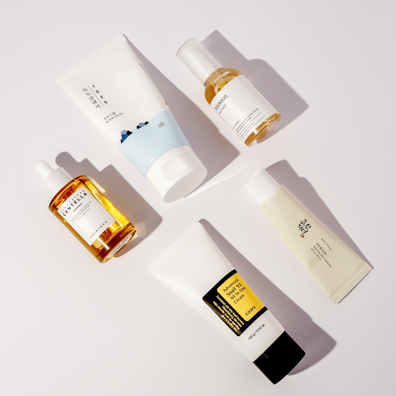 Flatlay von fünf K-Beauty Produkten: Round Lab 1025 Dokdo Creme, mixsoon Bean Essence, SKIN1004 Madagascar Centella Ampoule, COSRX Advanced Snail 92 All In One Cream und Beauty of Joseon Relief Sun Cream SPF50+ PA++++