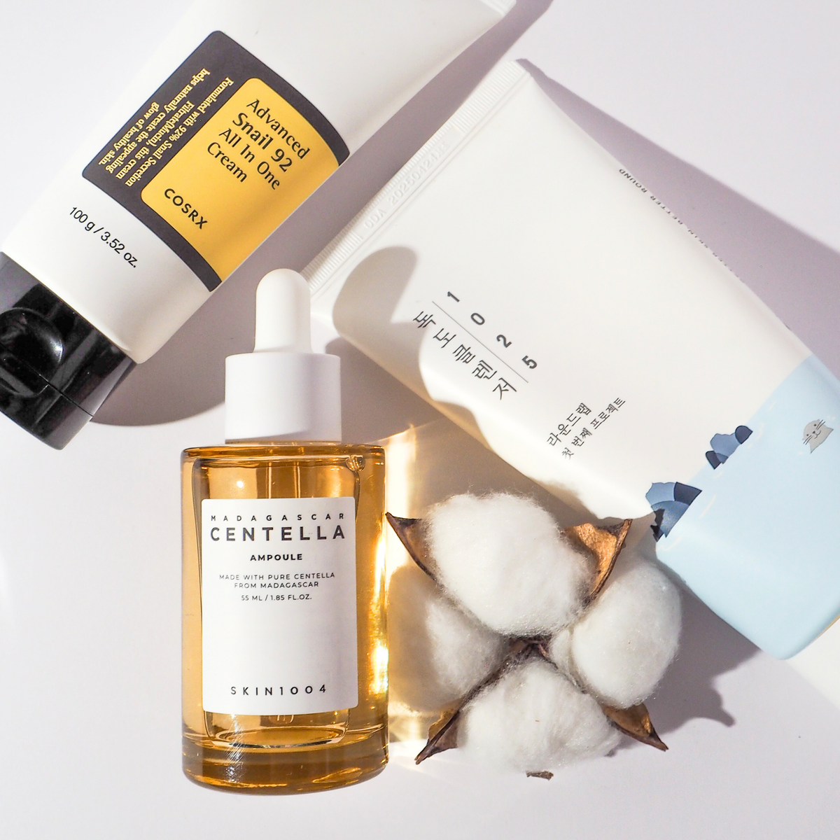 K-Beauty Flatlay mit COSRX Advanced Snail 92 All In One Cream, SKIN1004 Madagascar Centella Ampoule und Round Lab 1025 Dokdo Cleanser, dekoriert mit Baumwollkapseln
